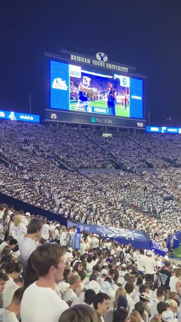 A packed whiteout crowd at LaVell Edwards Stadium watching Cosmo play Rock Paper Scissors on the videoboard, 8,681 fans in the round